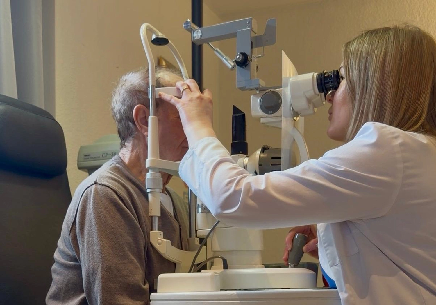 Praxis Olena Behrens – Augenheilkunde – Eine Frau untersucht das Auge einer Person mit einem Ophthalmoskop in einer klinischen Umgebung.