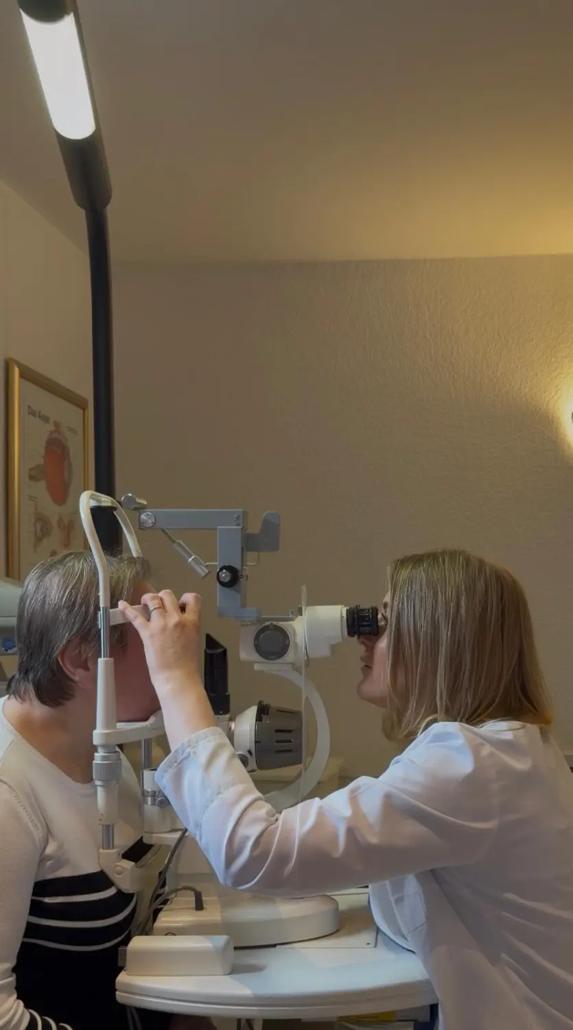 Praxis Olena Behrens – Augenheilkunde – Eine Frau untersucht ein Auge mit einem Ophthalmoskop in einer klinischen Umgebung.